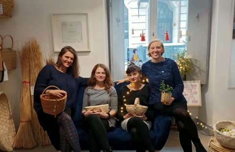 Na zdjęciu siedzą na granatowej kanapie 4 dziewczyny, każda z nich trzyma w rękach kosz wyplatany ręcznie, wokół nich znajdują się inne kosze i świąteczne światełka, zdjęcie świąteczne